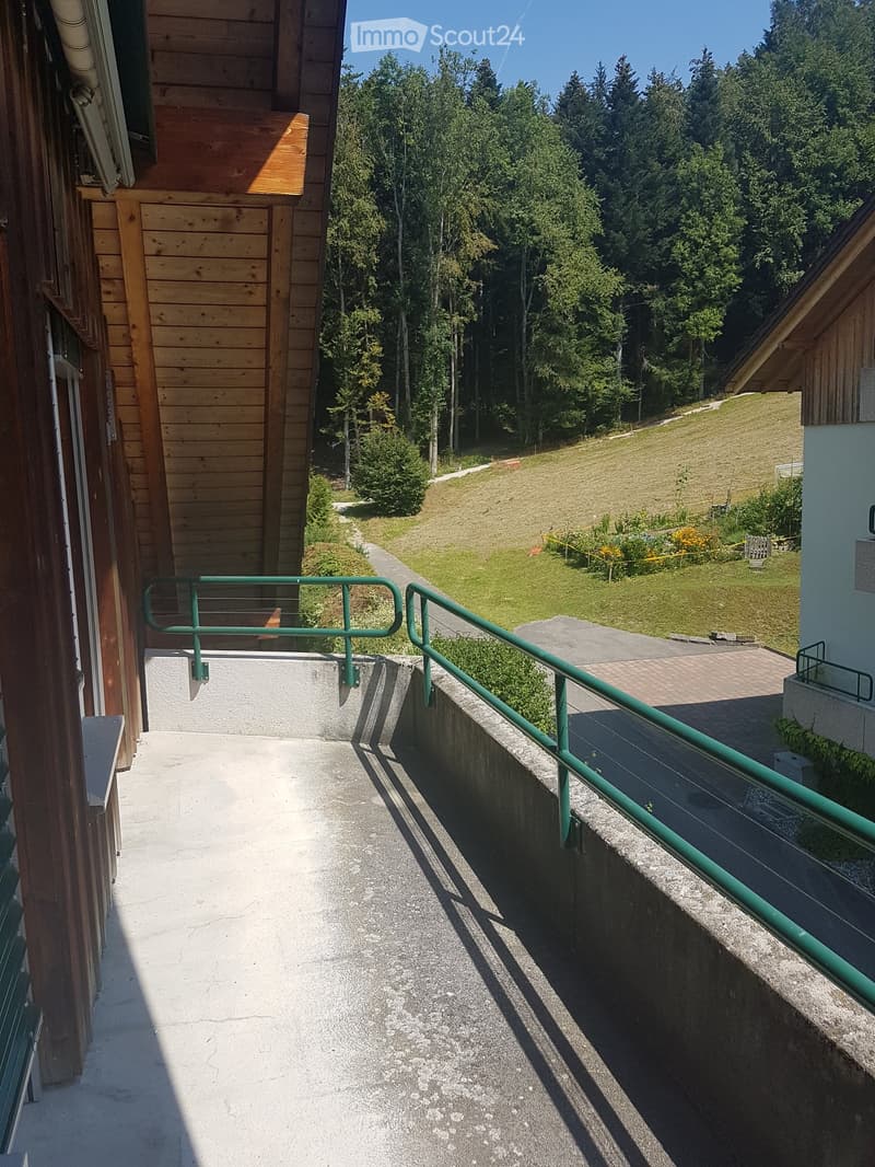 A narrow concrete pathway with green metal railings, leading to a grassy area surrounded by trees and a wooden building.