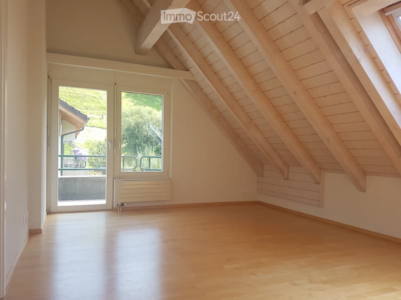 Empty attic room with wooden floors, exposed wooden beams, large windows, and a sliding glass door leading to a balcony.
