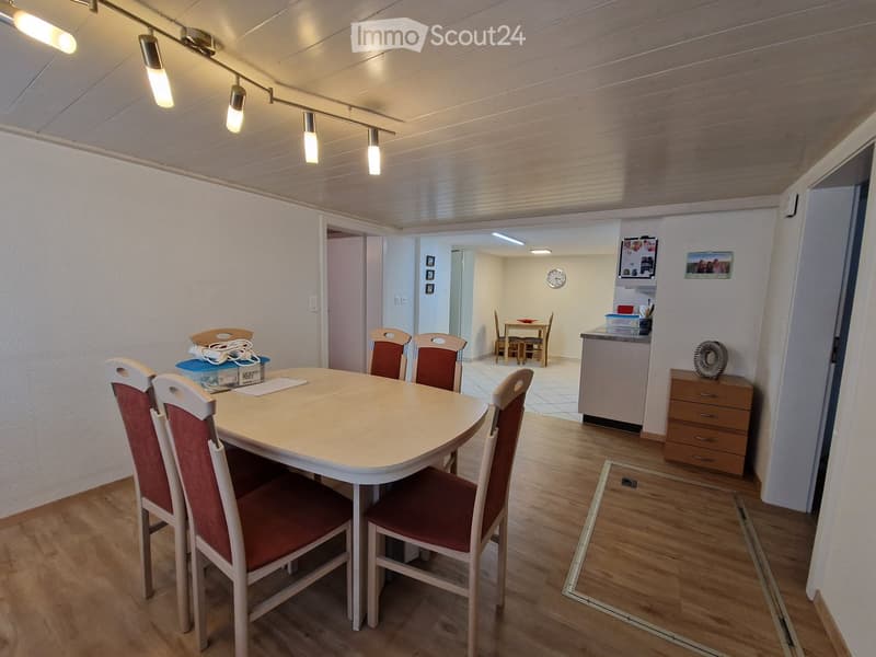 Dining area with a round table and six chairs, adjacent to a small kitchen area with a counter, cabinet, and clock on the wall.