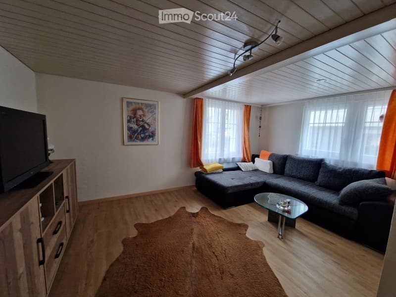 Modern living room with wooden flooring, grey sectional sofa, glass coffee table, TV stand, and artwork on the wall.