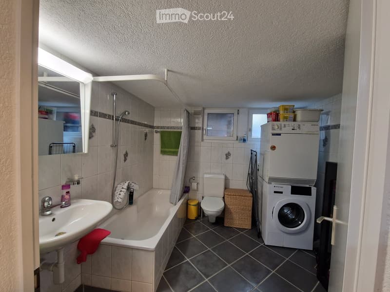 Bathroom with a sink, bathtub, toilet, washing machine, mirror, and towel. The room has tiled walls and a dark tiled floor.