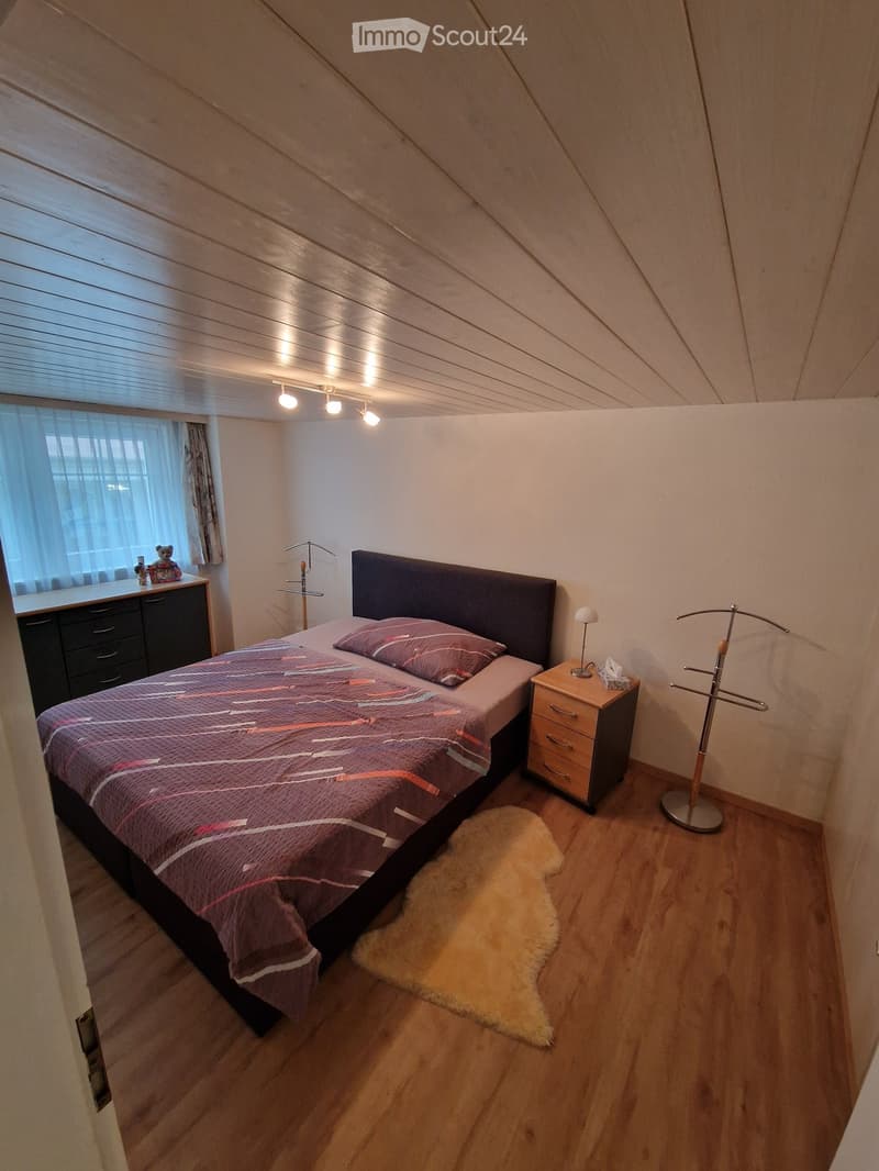 Cozy bedroom with wooden flooring, slanted ceiling, and modern decor. Features a bed with a dark headboard, patterned bedding, a nightstand with a lamp, a drawer unit, a fur rug, and a clothing rack.