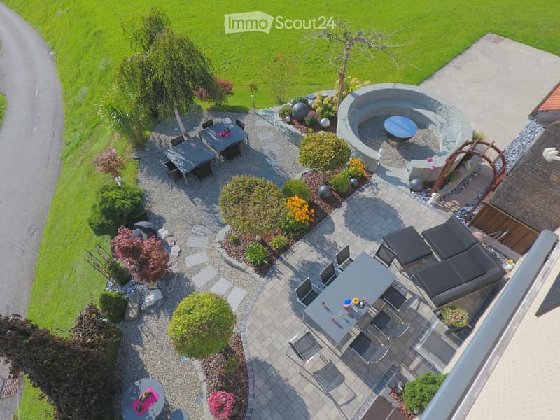 Well-maintained garden with a patio area, outdoor dining table, lounge chairs, and a circular fire pit surrounded by gravel and greenery.