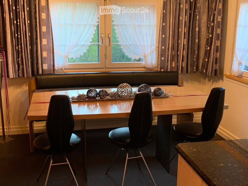 A small, cozy dining area with a wooden table, black chairs, and a built-in leather sofa bench. The room features large windows with curtains and a decorative centerpiece on the table.