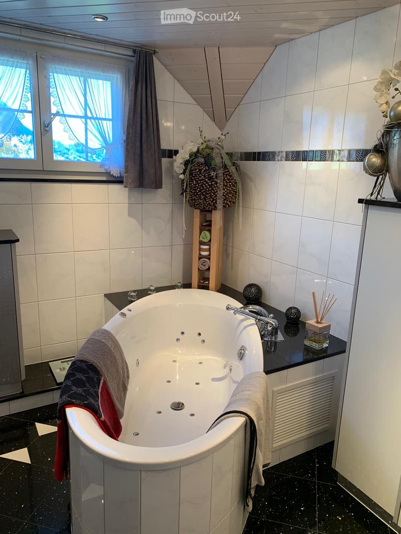 Modern bathroom with a large, oval bathtub, tiled walls, a window with curtains, and decorative items including a flower arrangement and a vase on the counter.