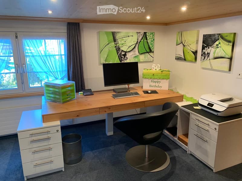 A modern, well-lit office space with a large wooden desk, ergonomic chair, computer setup, printer, and artwork on the walls.