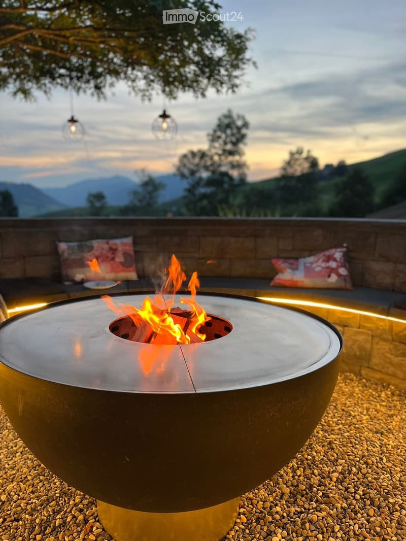 Outdoor patio with a modern fire pit surrounded by cushions, overlooking a scenic mountain view.