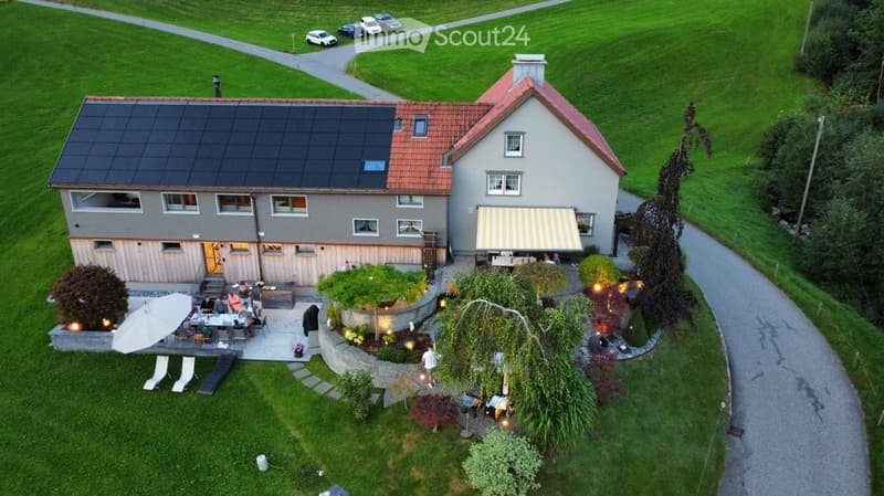 Aerial view of a two-story house with solar panels on the roof, surrounded by a well-maintained garden, patio area, and outdoor seating. The house features a garage, multiple windows, and a covered entrance.