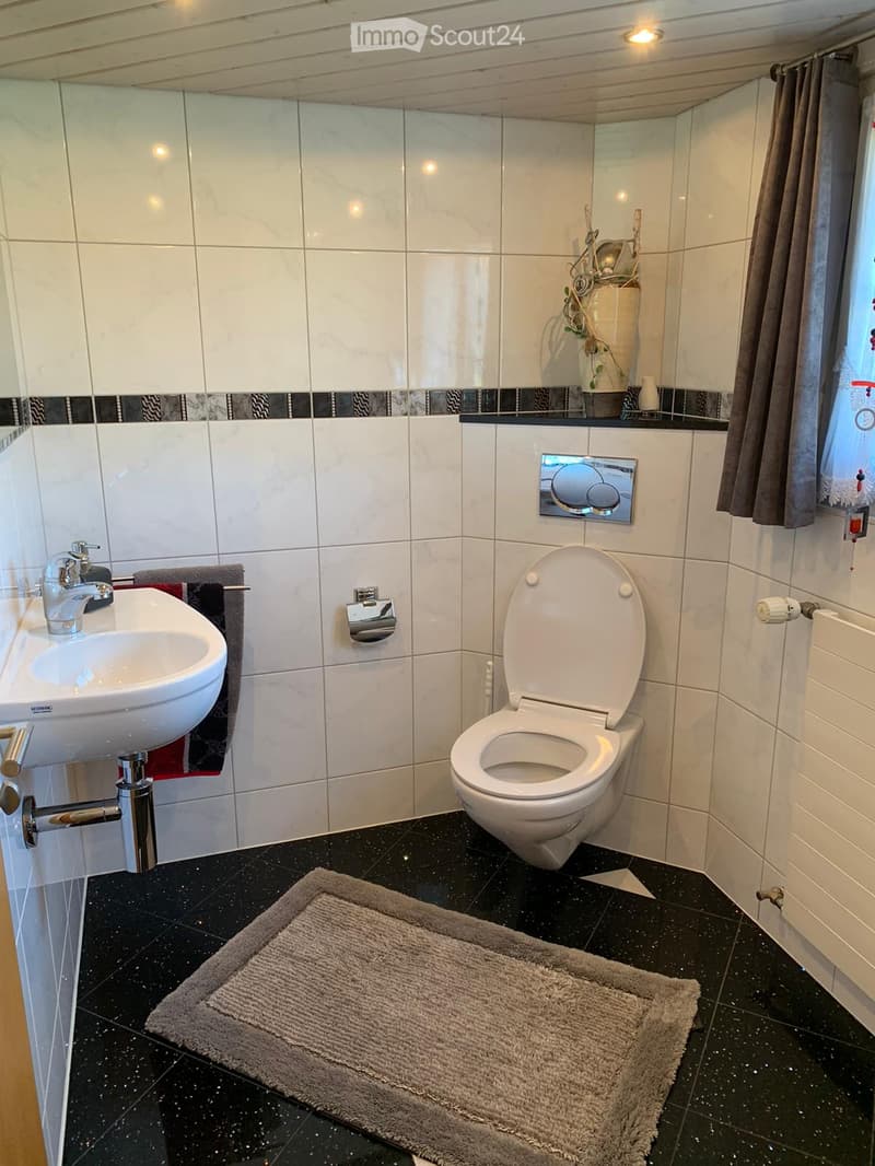 Modern bathroom with white tiles, black speckled floor, sink, toilet, towel, mirror, and a small rug.