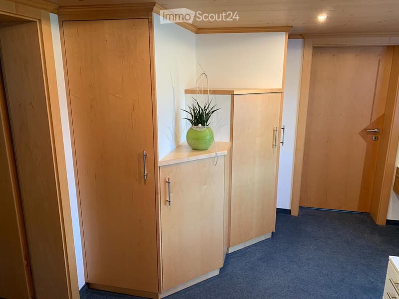 A narrow hallway with light wooden walls and doors, featuring a small shelf with a green vase and plant.