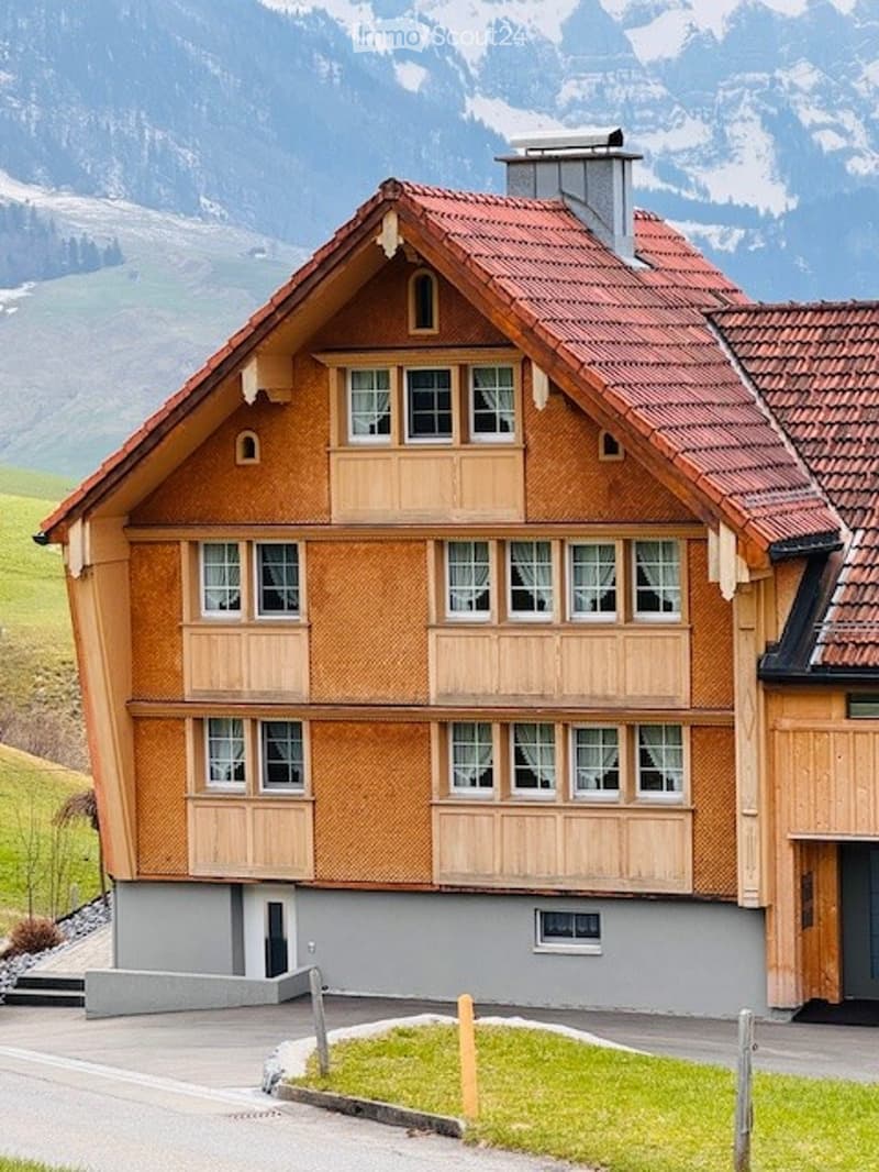 Two-story wooden house with red-tiled roof, multiple windows, and a chimney, set against a backdrop of snow-capped mountains.