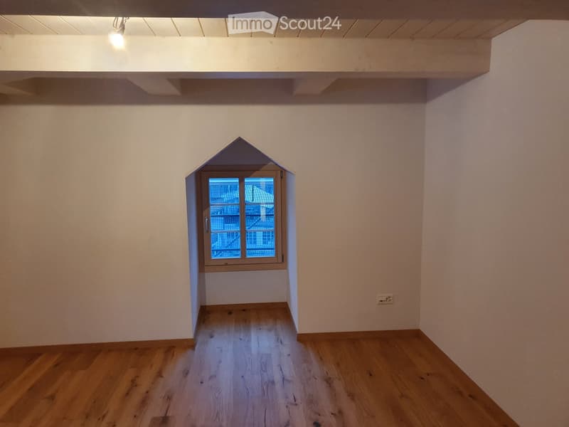 Empty room with wooden flooring, white walls, and a small window with a unique triangular frame.