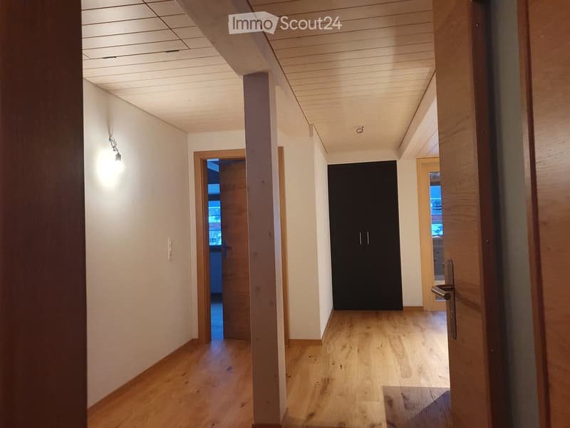 Empty hallway with wooden flooring, white walls, and recessed lighting. Features a black closet door and an open doorway leading to another room.