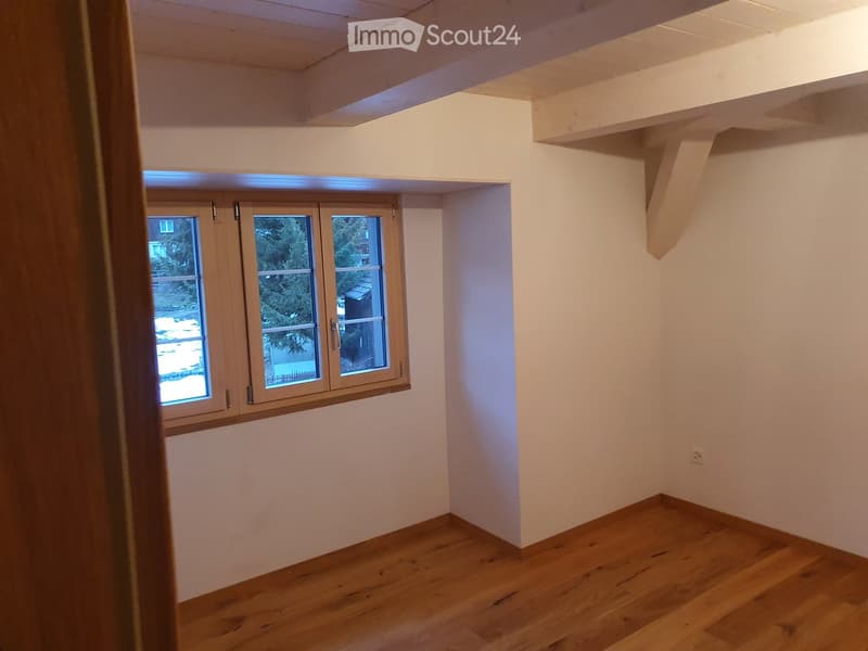 Empty room with wooden flooring, white walls, and a large window with wooden frame showing outdoor view.