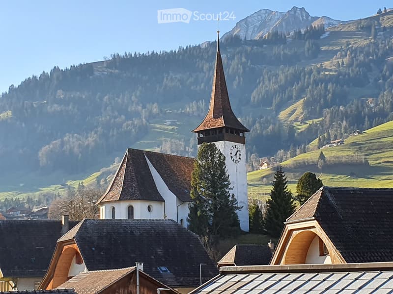 A picturesque village scene featuring a white church with a tall clock tower and pointed spire, surrounded by traditional wooden houses with dark roofs and surrounded by lush green hills and mountains.