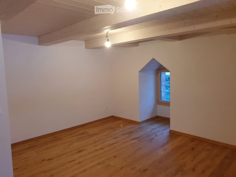 Empty room with wooden flooring, white walls, and a small arched doorway leading to another space with a window.
