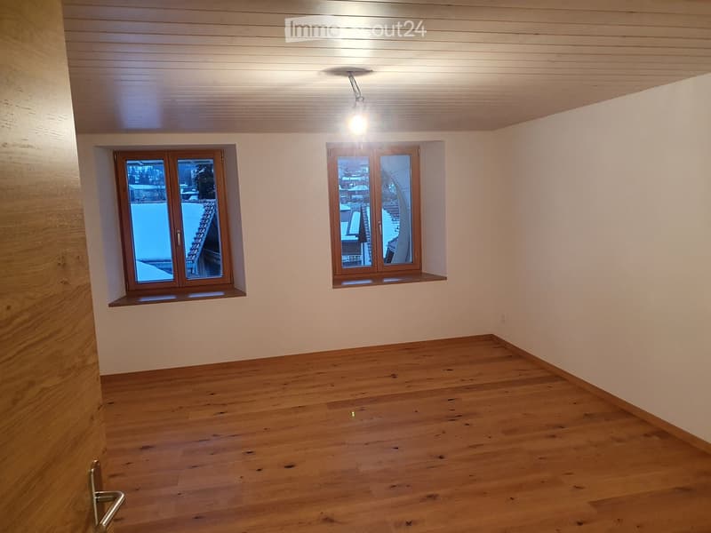 Empty room with wooden flooring, white walls, and two windows with wooden frames. The room has a simple, clean design with a single light fixture on the ceiling.
