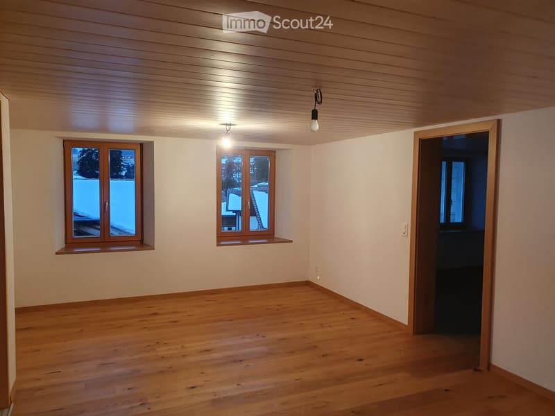 Empty room with wooden flooring, light-colored walls, and wooden ceiling. Two windows with wooden frames and a small shelf beneath one window. A doorway leads to another room.