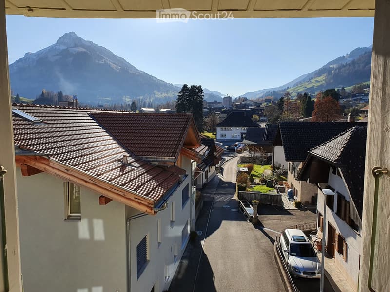 Balcony view overlooking a narrow street lined with traditional houses, with a scenic mountain backdrop and parked cars visible below.