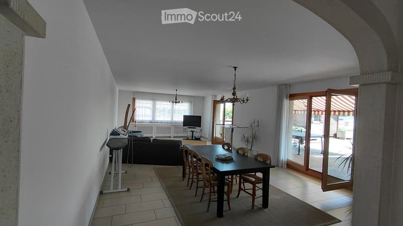 Open-plan living and dining area with a sofa, dining table, chairs, TV, chandelier, and exercise bike. Large windows and sliding glass doors lead to a balcony or terrace.