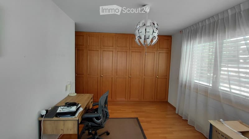 A well-lit room with wooden flooring, a large wooden cabinet, a modern hanging light fixture, a desk with a chair, and a lamp on the desk.