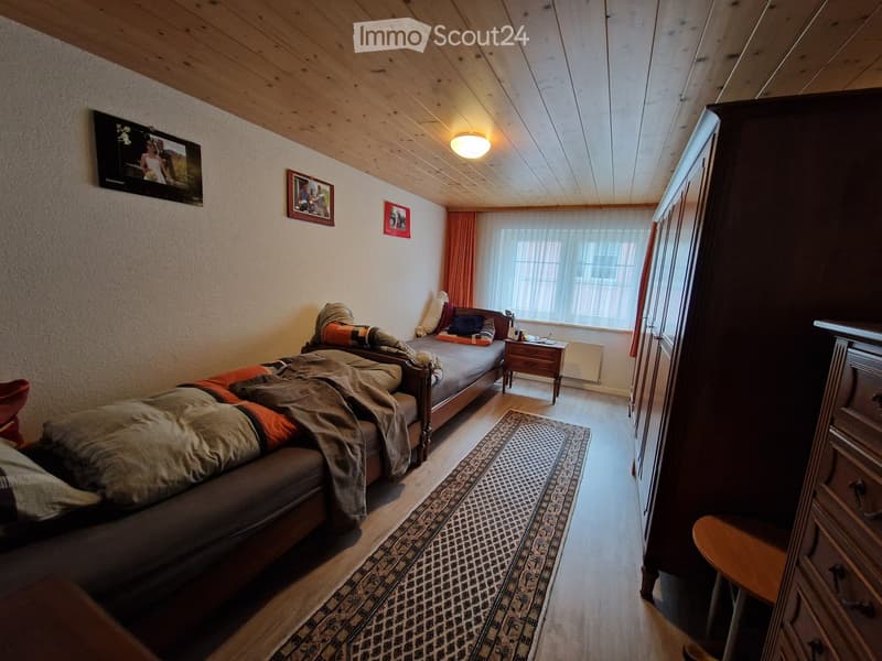 Cozy bedroom with wooden floors, slanted wooden ceiling, bed with pillows and blankets, nightstand, wardrobe, and television mounted on wall.