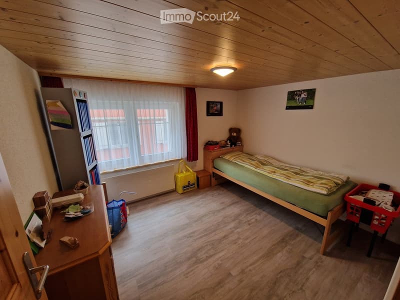 Cozy bedroom with wooden bed, nightstand, desk, and various personal items. Features wooden flooring, large window with curtains, and a slanted wooden ceiling.