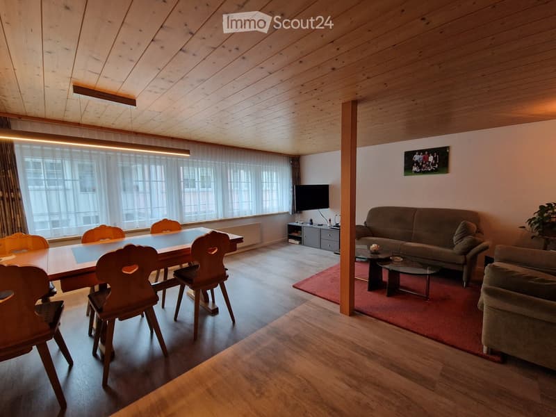 Spacious living and dining area with wooden floors, exposed wooden ceiling beams, large windows with sheer curtains, a sofa, a coffee table, a dining table with chairs, and a television on a stand.