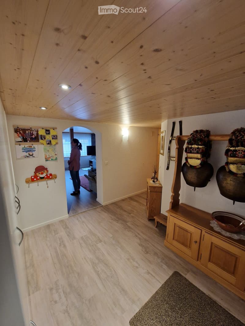 Wooden-floored hallway with warm lighting, a small wooden cabinet, decorative items, and an archway leading to another room.