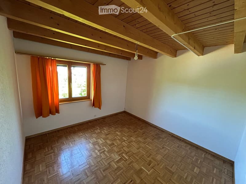Empty room with wooden parquet flooring, white walls, and exposed wooden ceiling beams. A single window with orange curtains allows natural light to enter.