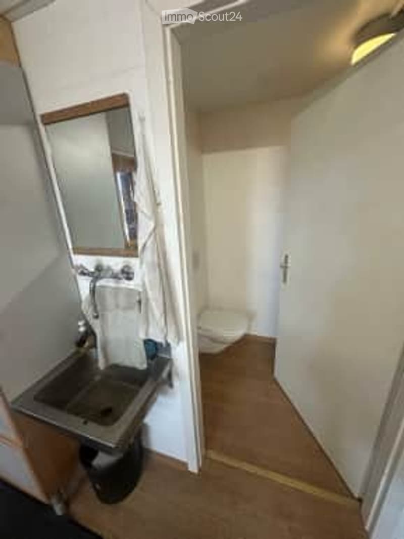 Compact bathroom with a sink, mirror, and toilet. The space is clean and functional with light-colored walls and wooden flooring.