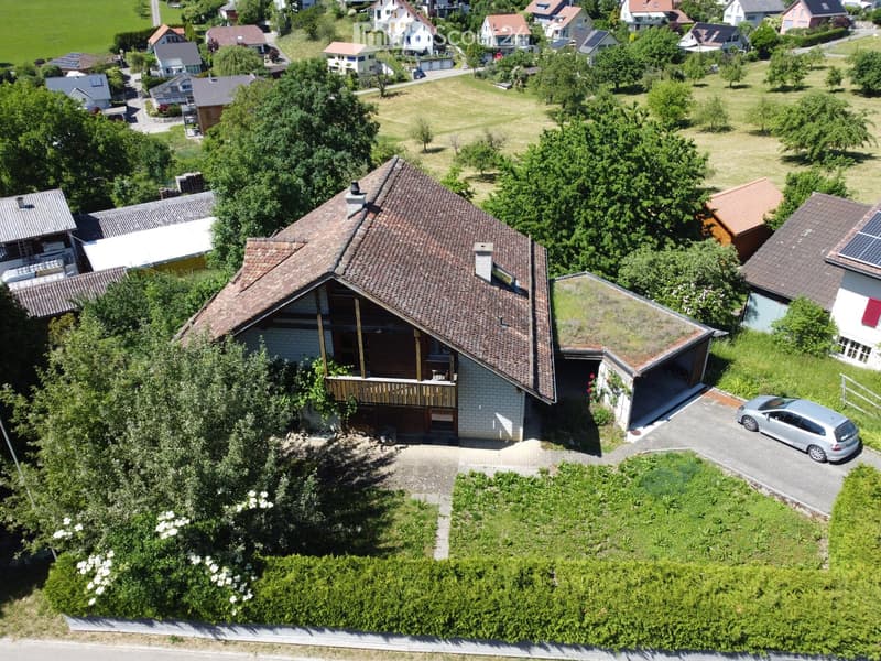 Aerial view of a two-story house with a brown tiled roof, a wooden balcony, and a driveway with a parked car. The house is surrounded by greenery and trees, with neighboring houses visible in the background.