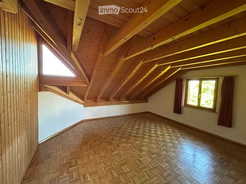 Empty attic room with wooden slanted ceilings, wooden flooring, and two windows with curtains.