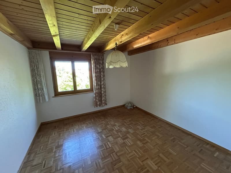 Empty room with wooden parquet flooring, white walls, exposed wooden ceiling beams, and a single window with curtains. A hanging lamp is visible.