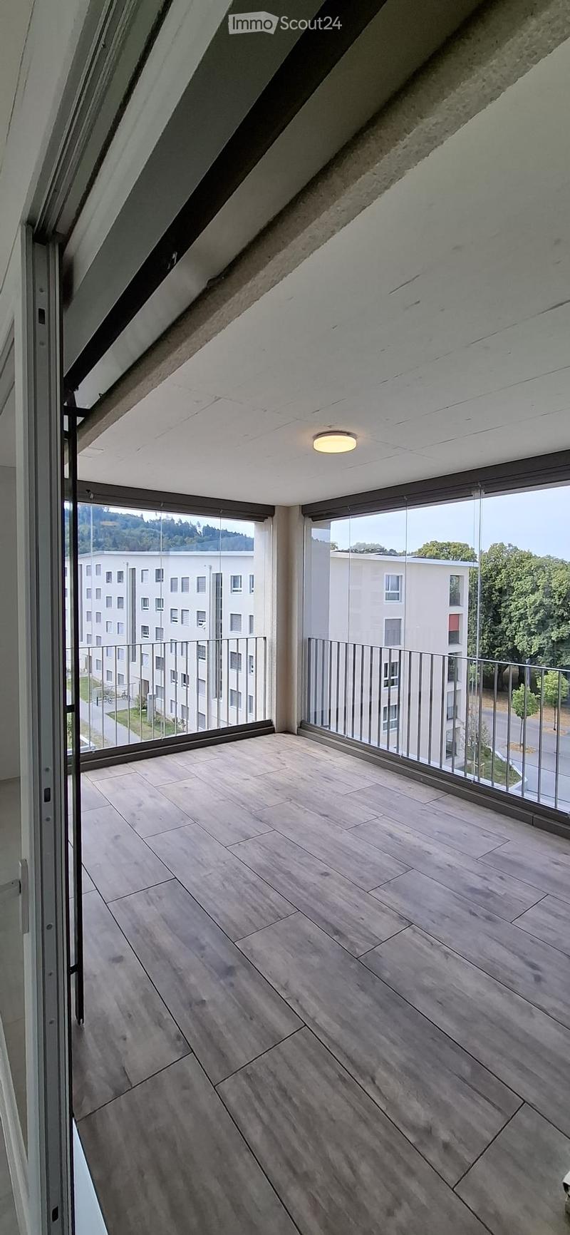 Modern balcony with wooden flooring, large glass windows, and metal railings, offering a view of surrounding buildings and greenery.