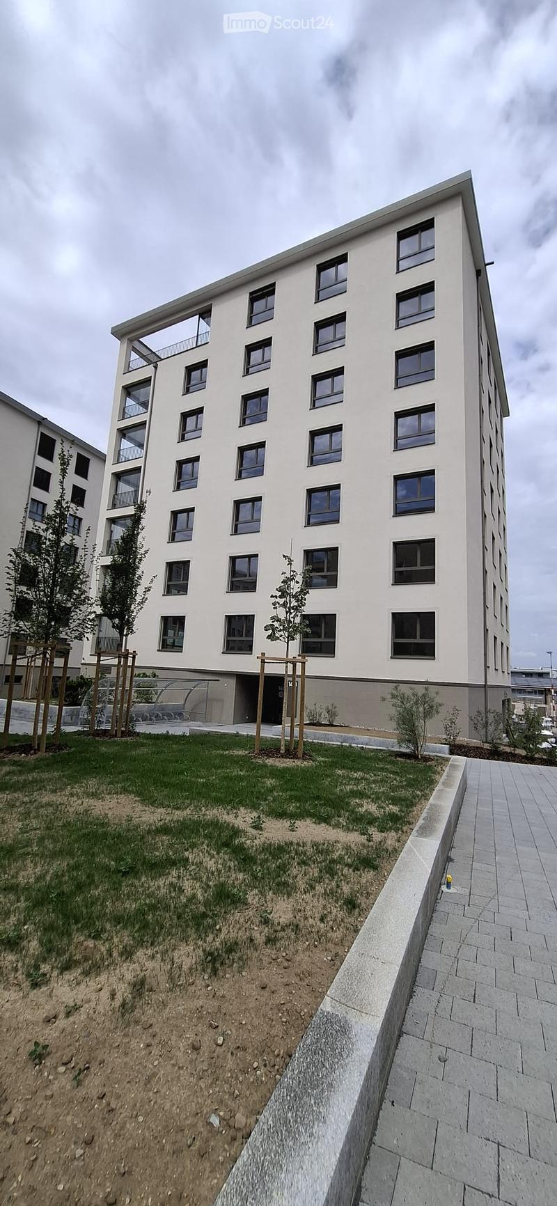 Modern multi-story residential building with white facade, multiple windows, and balconies. The ground level features a landscaped area with grass, small trees, and a paved walkway.