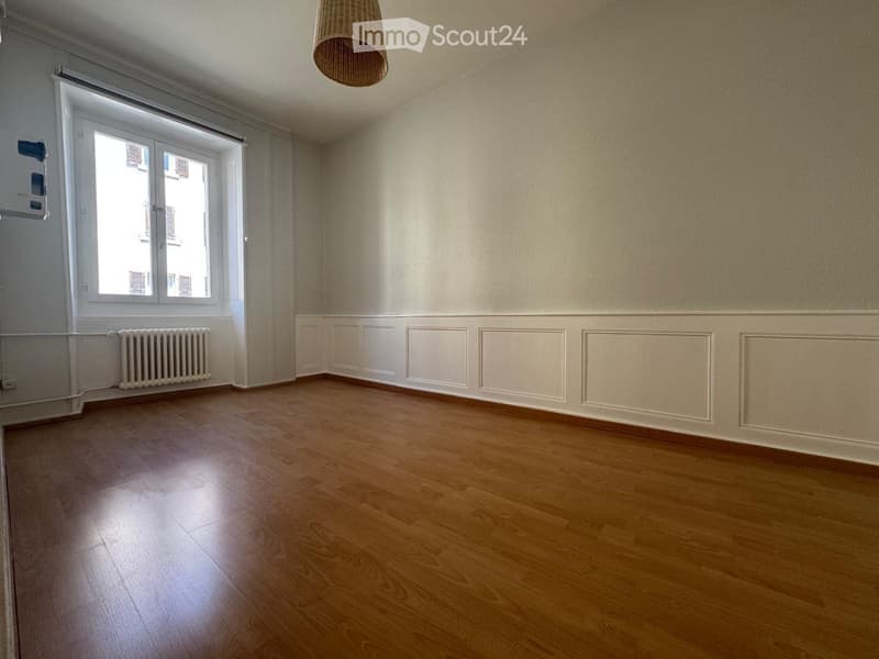 Empty room with wooden flooring, white walls, large window, radiator, and a hanging light fixture.