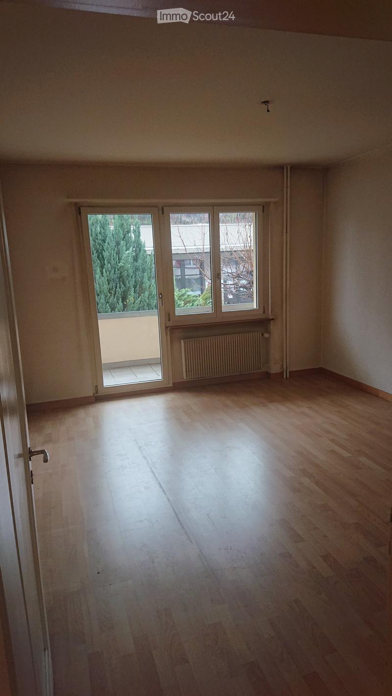 Empty room with wooden flooring, large window with sliding doors leading to a balcony, radiator, and visible light fixture on the wall.