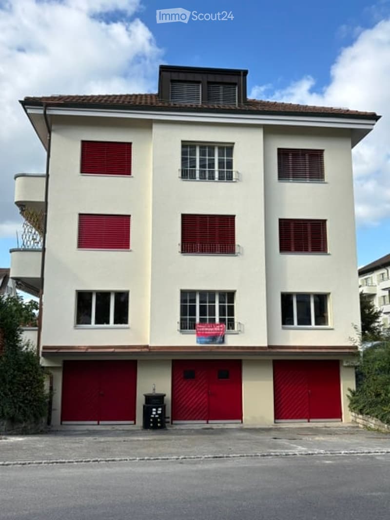 Three-story residential building with beige facade, red shutters, and three garage doors at the ground level.