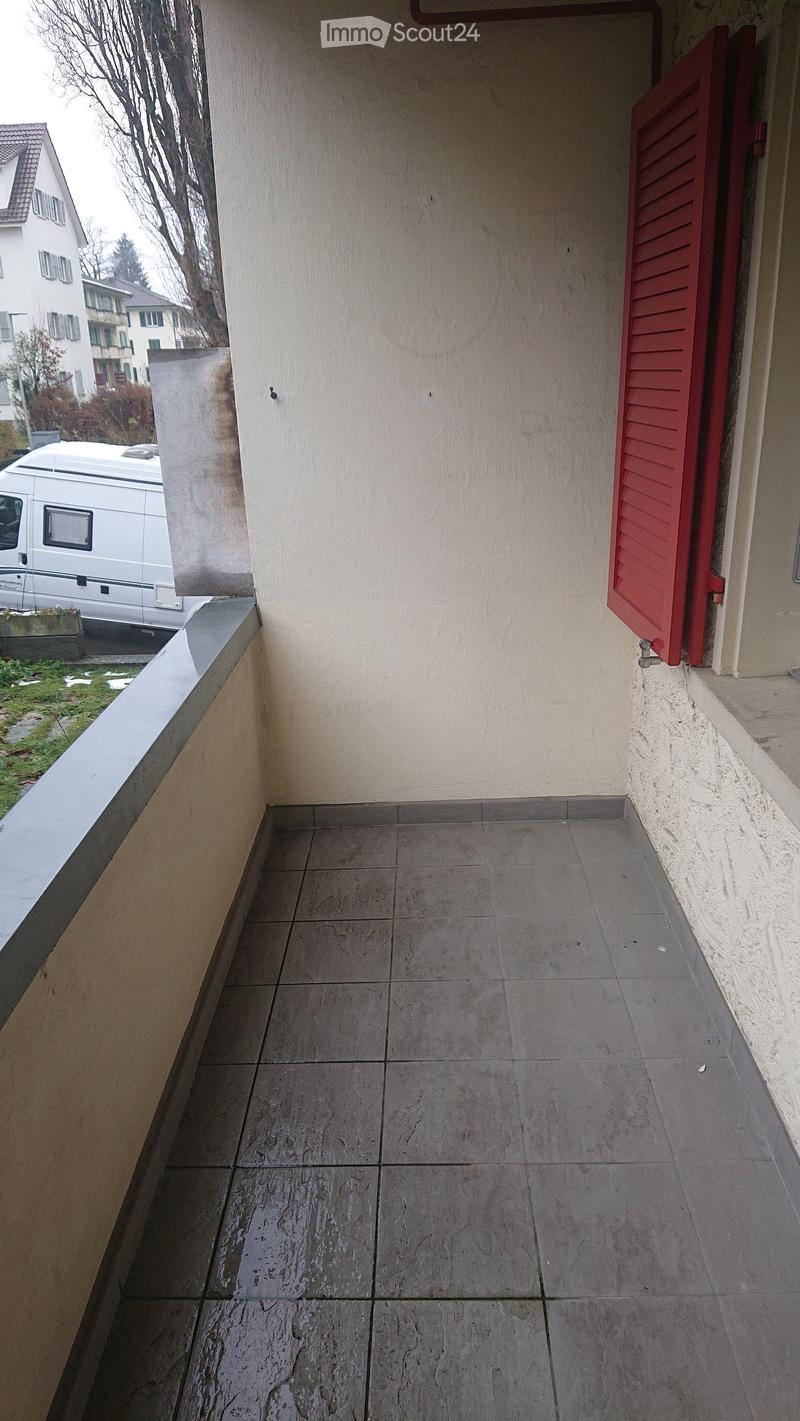 Balcony with tiled floor, beige walls, and red shutters on the right side. The balcony overlooks a grassy area with a parked van and neighboring buildings.