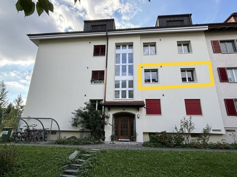 Four-story residential building with white facade, large windows, and red shutters. Features a central entrance with a wooden door, small garden area, and bicycles parked near the entrance.