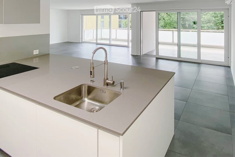 Modern kitchen with white cabinets, light grey countertops, a stainless steel sink, and a sleek faucet. Large windows and sliding glass doors lead to a balcony.