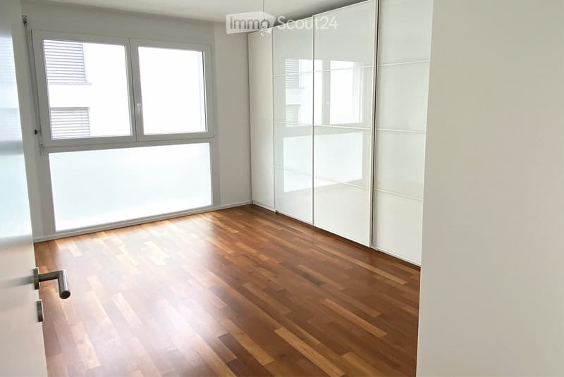 Empty room with wooden flooring, white walls, large windows, and a sliding wardrobe unit.