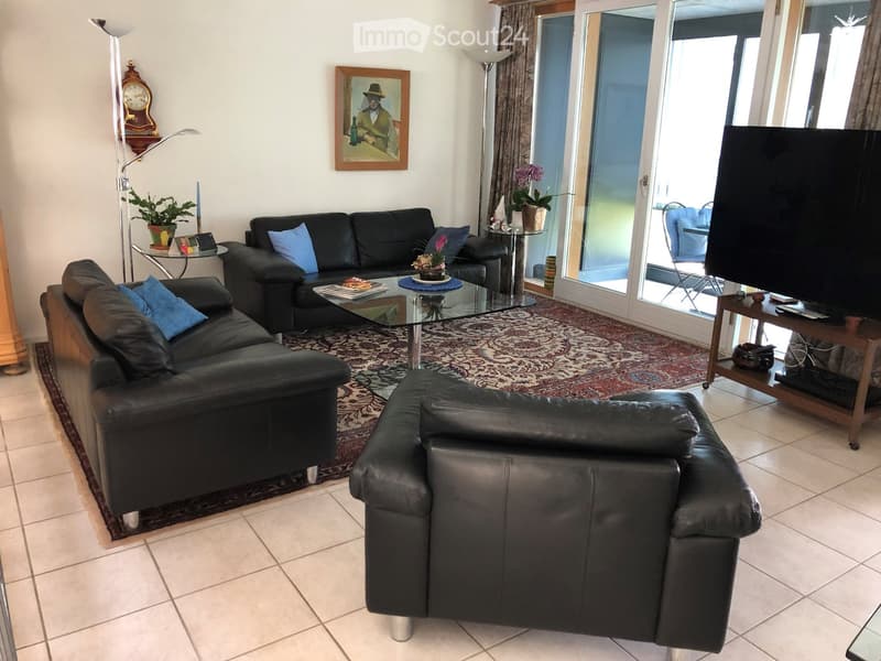 Modern living room with black leather sofa, armchair, coffee table, TV stand, floor lamp, and a view through large glass doors to a patio area.