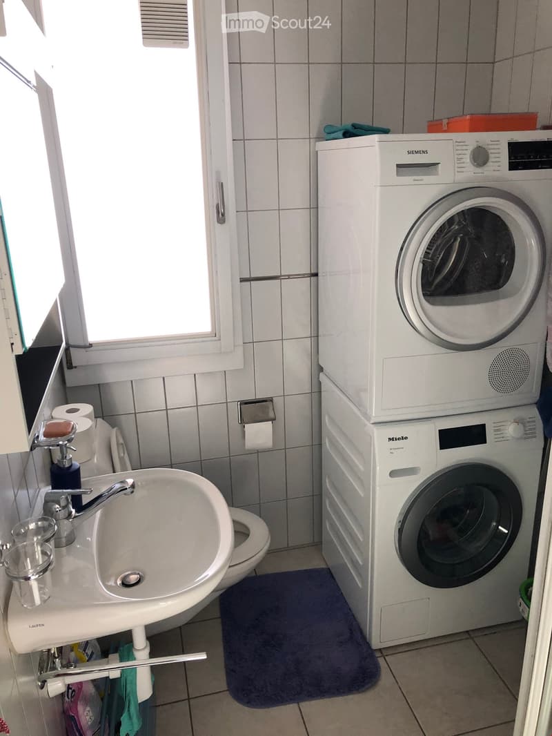 Bathroom with a white sink, faucet, toilet, and stacked washing machine and dryer unit. Includes a soap dispenser, toilet paper, and a cup on the sink.