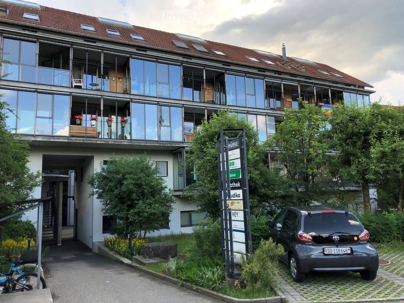 Modern multi-story building with large glass windows and balconies, featuring a small car parked in front and a bicycle on the left side.
