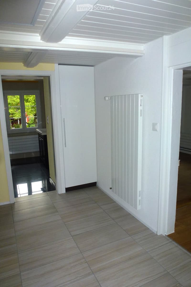 Empty hallway with tiled floor, white walls, radiator, and a door leading to another room.