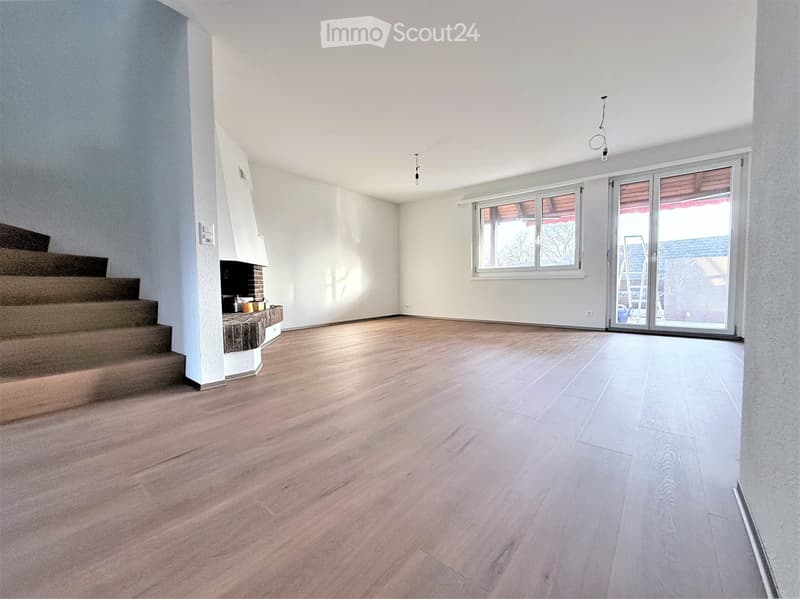 Empty room with wooden flooring, white walls, and a staircase leading upwards. A fireplace is visible on the left side, and a large window with a sliding door is on the right.