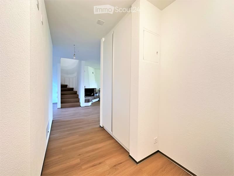 A narrow hallway with light wooden flooring, white walls, and a ceiling light fixture. The hallway leads to a staircase and an open doorway to another room.