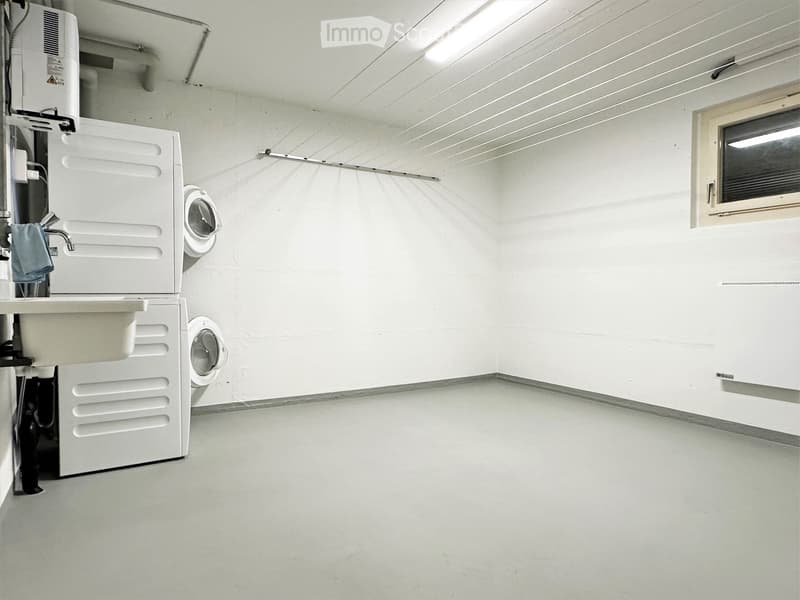 A small, clean bathroom with white walls and a concrete floor. It features a washing machine, a dishwasher, and a sink with a faucet.